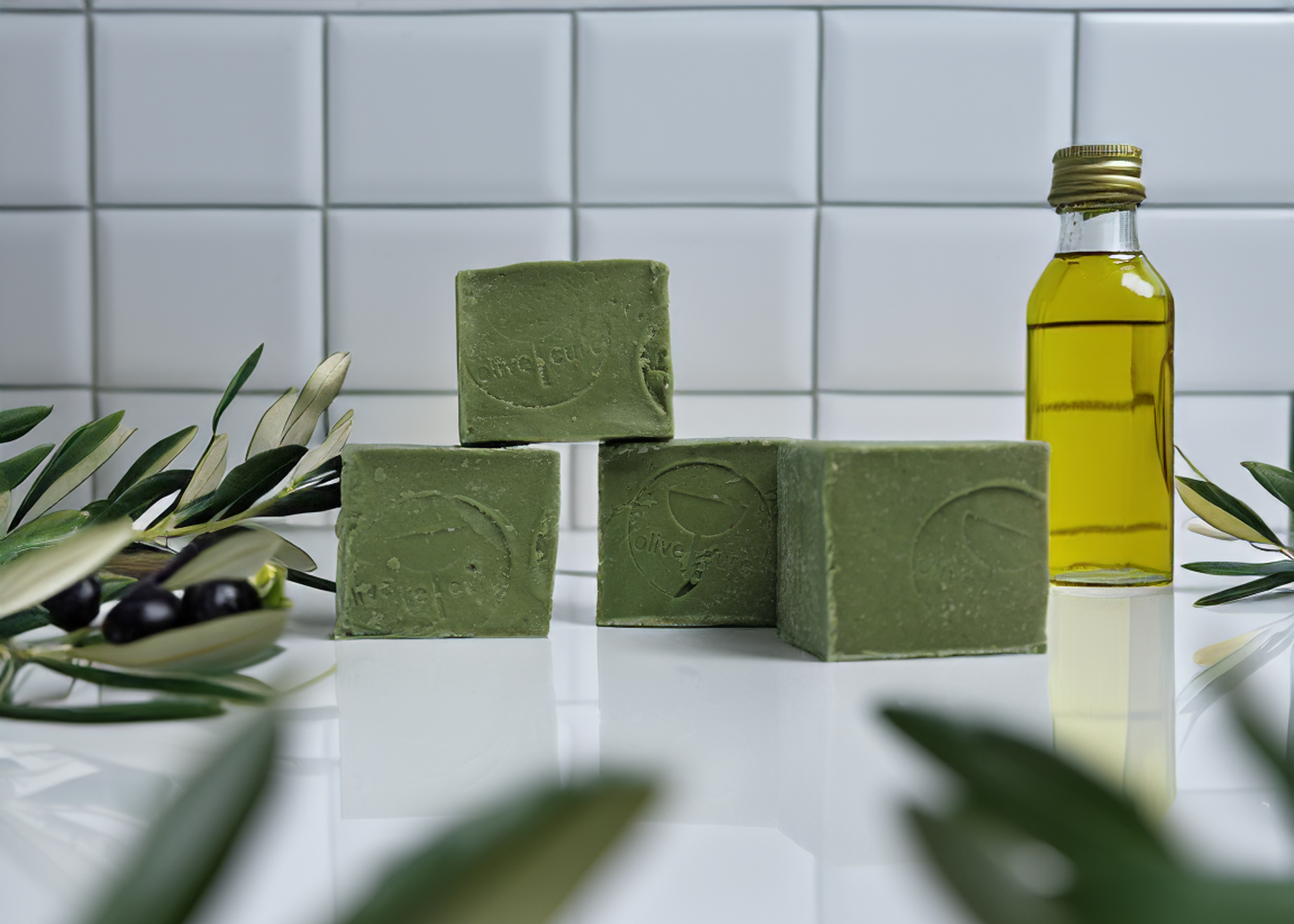 a clearn white counter top with olive branches black olives and olive oil around four bars of ghar soap made from olive oil and ghar oil by nablus soap company, known as olive cure, skin care soap