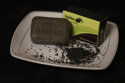 Black cumin and olive oil soap bar by nablus soap company, the black soap bar is sitting on a white soap tray with the box next to it and a pile of fresh black cumin showcasing the ingredients in the soap bar